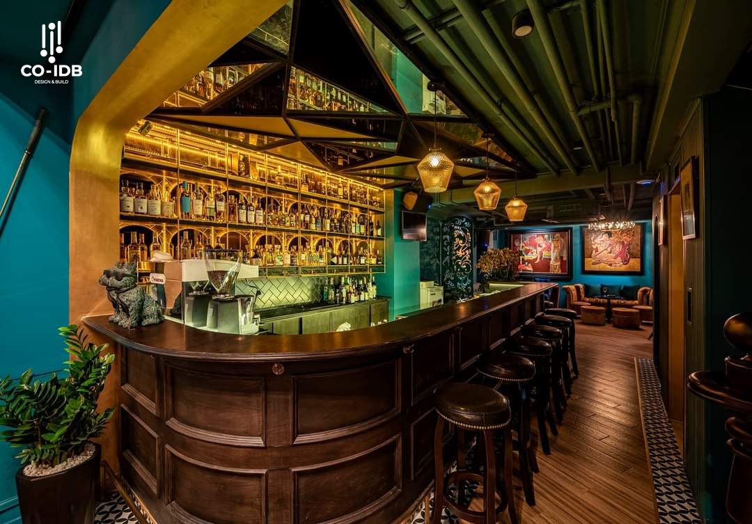 Quầy bar nội thất gỗ đậm chất Đông Dương, phối hợp đèn trần ánh vàng cùng trần sơn xanh cổ vị, điểm nhấn gương kính phản chiếu tạo chiều sâu và hiệu ứng ánh sáng ấm áp.