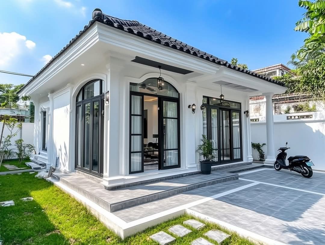 Mẫu nhà cấp 4 tân cổ điển mái Nhật màu trắng sang trọng, thiết kế cửa vòm kính khung đen nổi bật, có sân lát gạch xám và lối đi thảm cỏ xanh mướt.