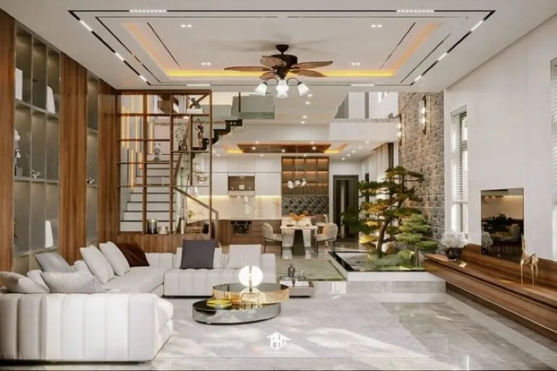 Phòng khách nhà phố hiện đại không gian mở, nổi bật với bộ sofa chữ L màu trắng, bàn trà tròn mặt kính mạ vàng, vách tường đá và tiểu cảnh cây xanh trong nhà.