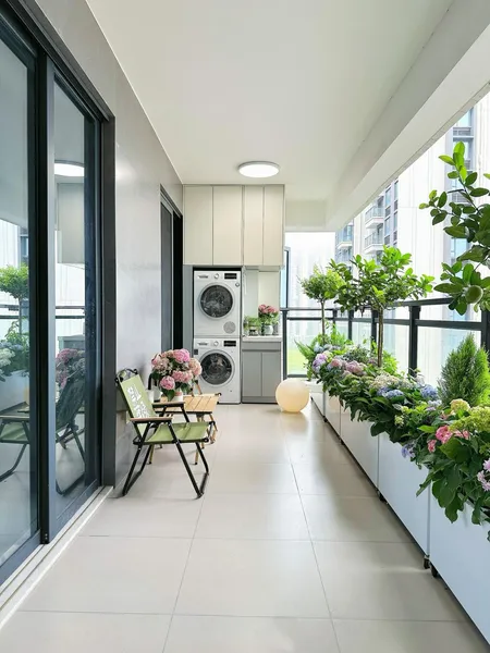 Không gian ban công chung cư rộng rãi với bộ máy giặt sấy chồng tầng gọn gàng ở góc, hệ thống bồn cây hoa cẩm tú cầu dọc lan can kính và một góc thư giãn nhỏ đặt ghế xếp.