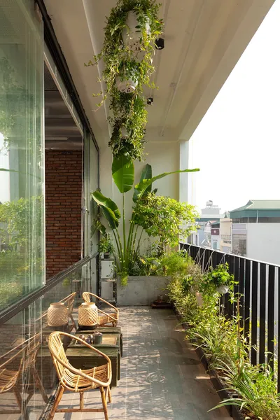 Không gian ban công dài với lan can sắt dọc, sàn đá, bộ bàn ghế mây tre đan nhỏ gọn, chậu cây dây leo treo thả từ trần, cây chuối cảnh lớn ở góc và bồn cây xanh chạy dọc lan can.