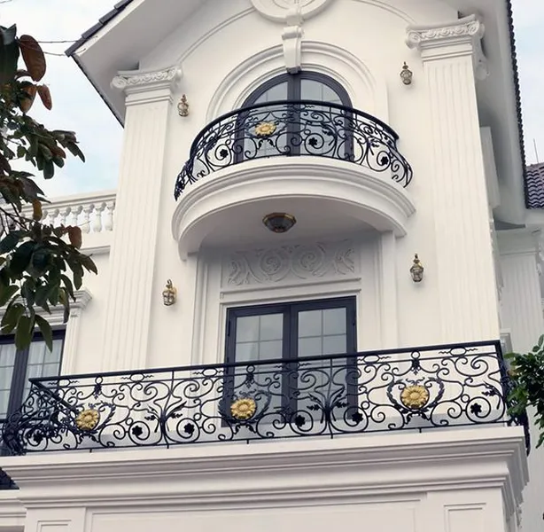 Mặt tiền biệt thự phong cách tân cổ điển màu trắng với hệ cột phào chỉ, cửa vòm và hai tầng ban công sở hữu lan can sắt mỹ thuật uốn hoa văn tinh xảo điểm hoa đồng mạ vàng sang trọng.